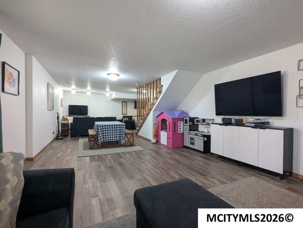 Basement living area with a large wall-mounted TV, white storage cabinet, and a pink children’s play kitchen next to a kid-sized playhouse. A table with a blue‑and‑white checkered cloth sits center-left, with seating around the room.