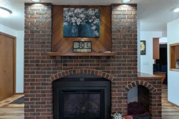 Brick fireplace with a wooden mantel, floral artwork above, and an arched brick alcove on the right; a pet bed and ball sit nearby.