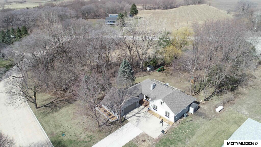 Aerial view of a residential area with a white house, driveway, and leafless trees in a large yard; a green tractor sits near a shed surrounded by trees.