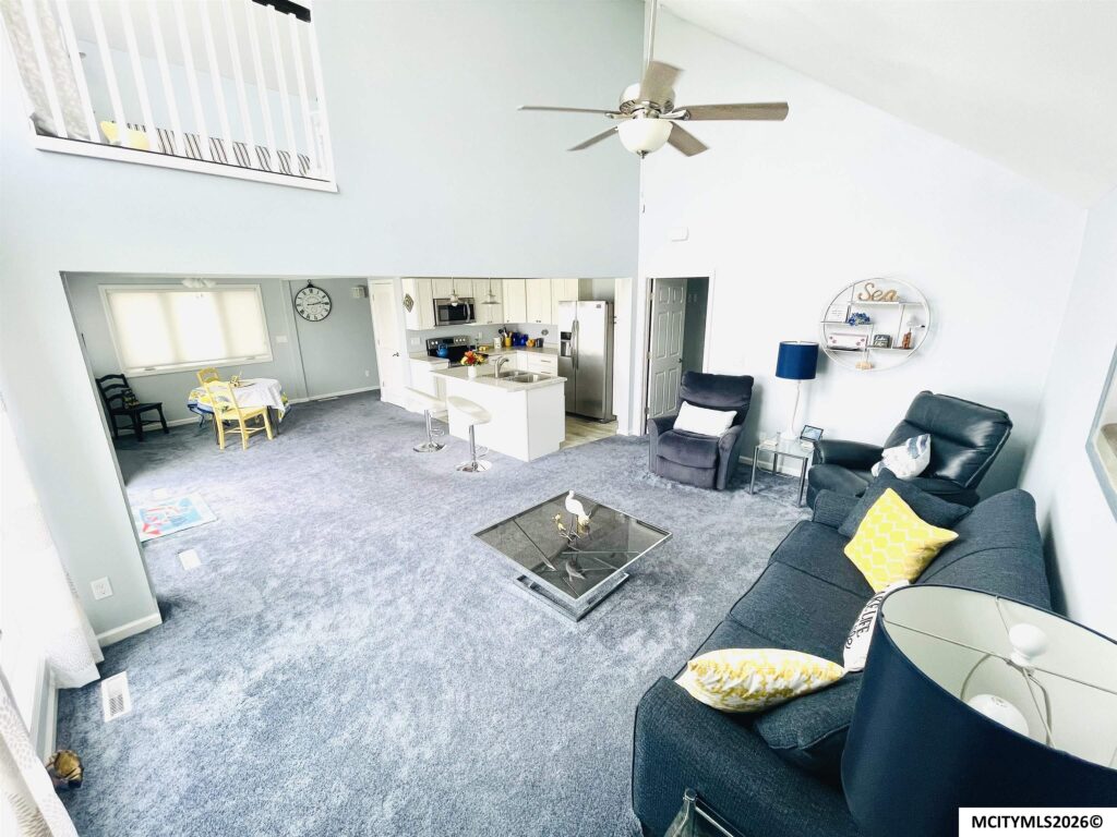 Open-concept living room with gray carpeting, blue walls, white kitchen, ceiling fan, and a round wall shelf with decor on the right."decorative round wall shelf with sea-themed items visible"