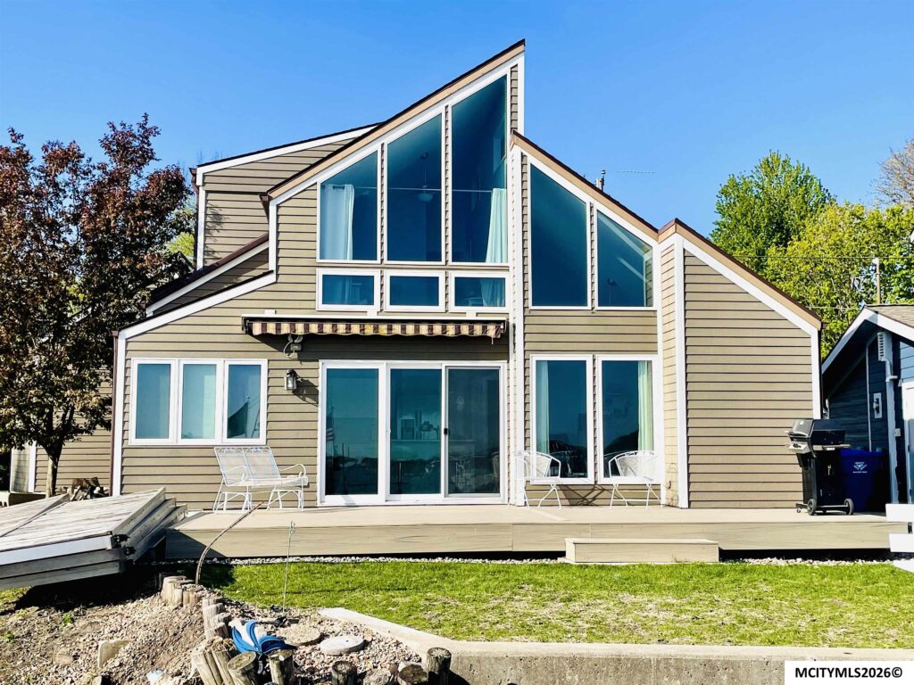 Contemporary house with expansive triangular glass walls and beige siding, set on a bright sunny day with patio chairs on a deck.